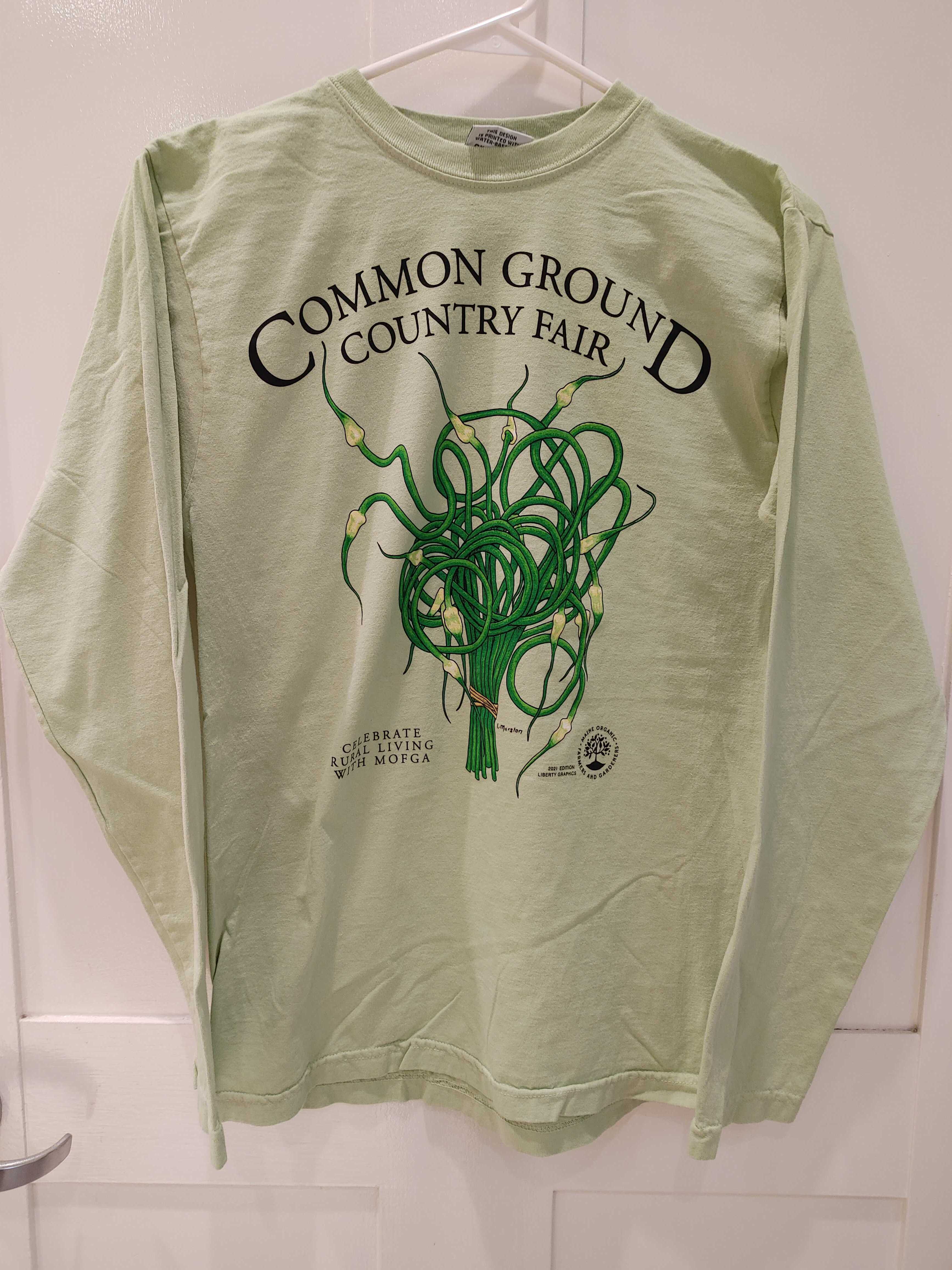 A light brown long sleeved shirt with artwork in the center depicting a cluster of green and cream garlic scapes bound with brown twine at the bottom. Black text around art reads “Common Ground Country Fair” and  “Celebrate Rural Living with MOFGA”. A black MOFGA logo is in the bottom right corner. 