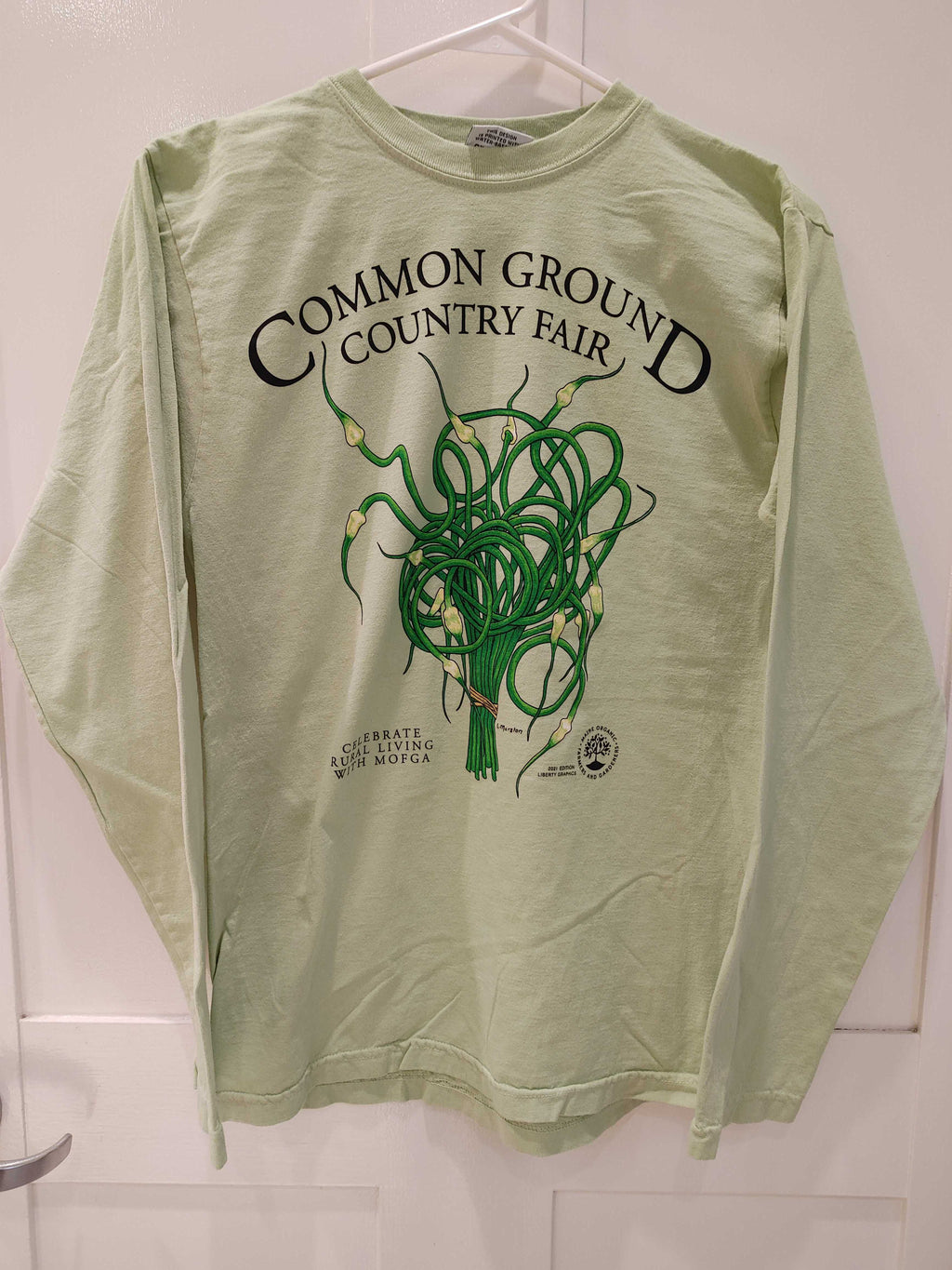 A light brown long sleeved shirt with artwork in the center depicting a cluster of green and cream garlic scapes bound with brown twine at the bottom. Black text around art reads “Common Ground Country Fair” and  “Celebrate Rural Living with MOFGA”. A black MOFGA logo is in the bottom right corner. 