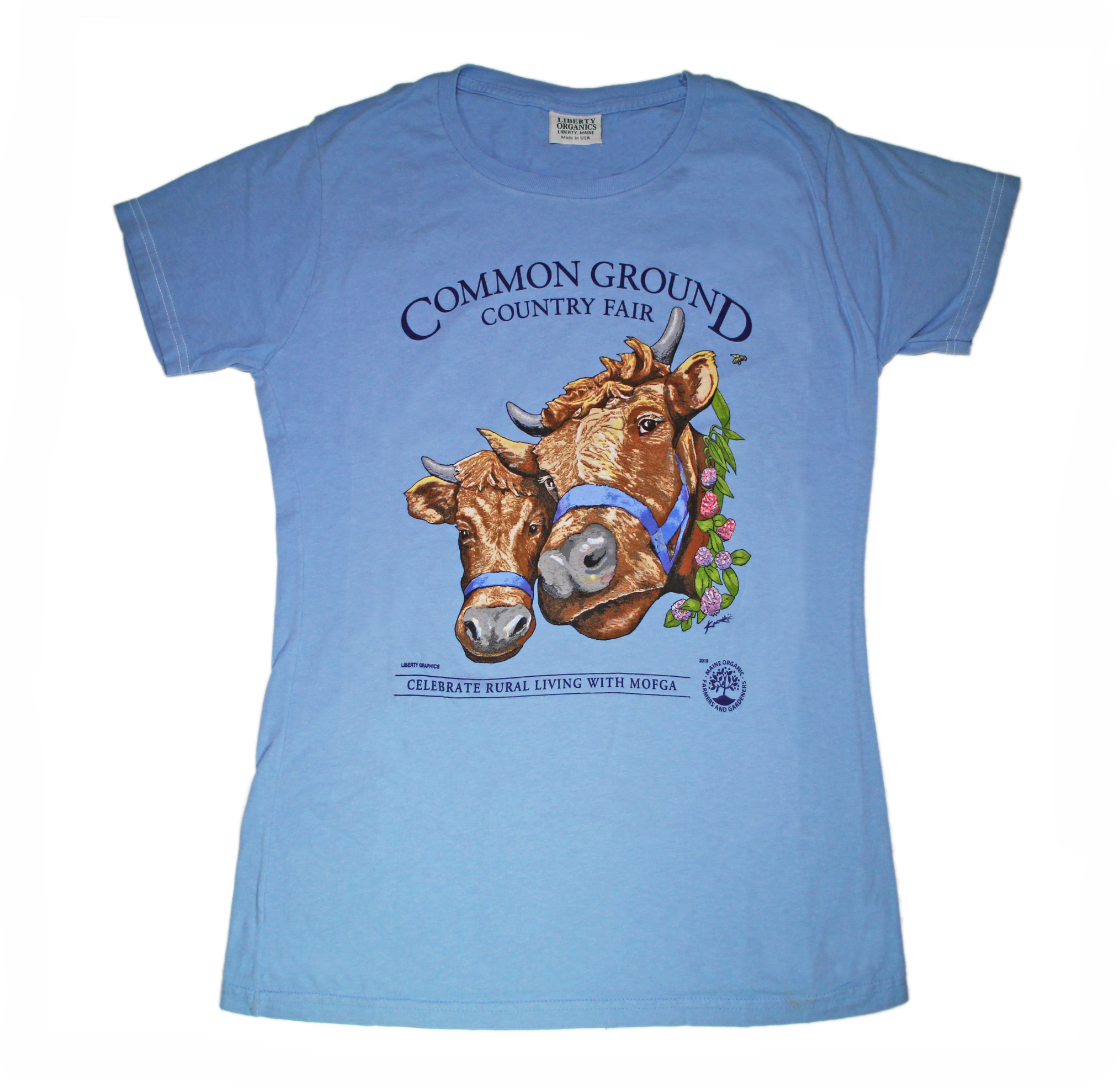 A sky blue shirt with artwork in the center depicting the heads of two shaggy brown cows with horns, wearing blue halters. On the right side of one cow is a viney wreath with small clusters of purple and blush flowers. Black text around art reads “Common Ground Country Fair '' and  “Celebrate Rural Living with MOFGA”. A black MOFGA logo is in the bottom right corner. 