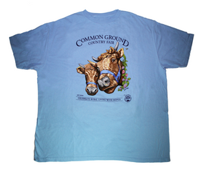A sky blue shirt with artwork in the center depicting the heads of two shaggy brown cows with horns, wearing blue halters. On the right side of one cow is a viney wreath with small clusters of purple and blush flowers. Black text around art reads “Common Ground Country Fair '' and  “Celebrate Rural Living with MOFGA”. A black MOFGA logo is in the bottom right corner. 