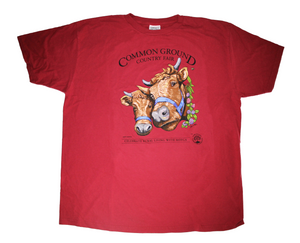 A red shirt with artwork in the center depicting the heads of two shaggy brown cows with horns, wearing blue halters. On the right side of one cow is a viney wreath with small clusters of purple and blush flowers. Black text around art reads “Common Ground Country Fair '' and  “Celebrate Rural Living with MOFGA”. A black MOFGA logo is in the bottom right corner. 