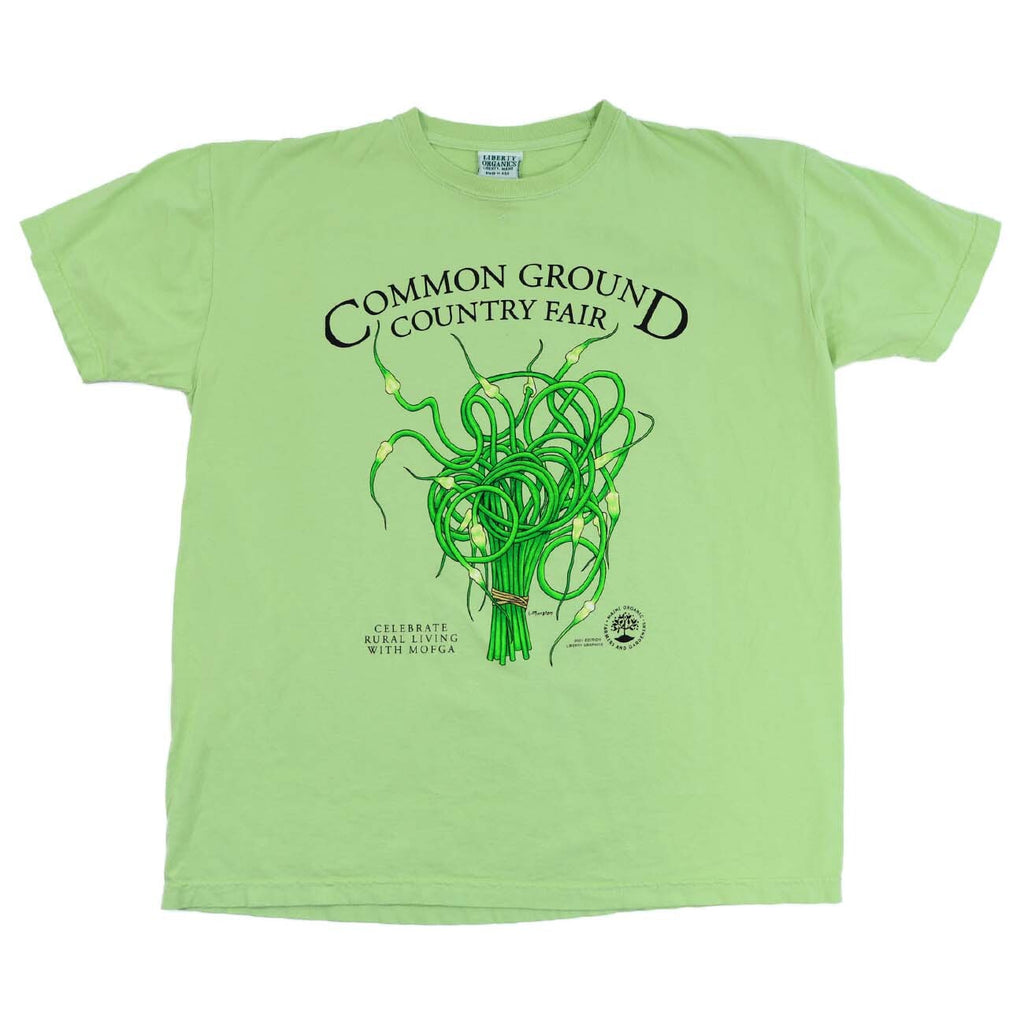 A light green shirt with artwork in the center depicting a cluster of green and cream garlic scapes bound with brown twine at the bottom. Black text around art reads “Common Ground Country Fair” and  “Celebrate Rural Living with MOFGA”. A black MOFGA logo is in the bottom right corner. 