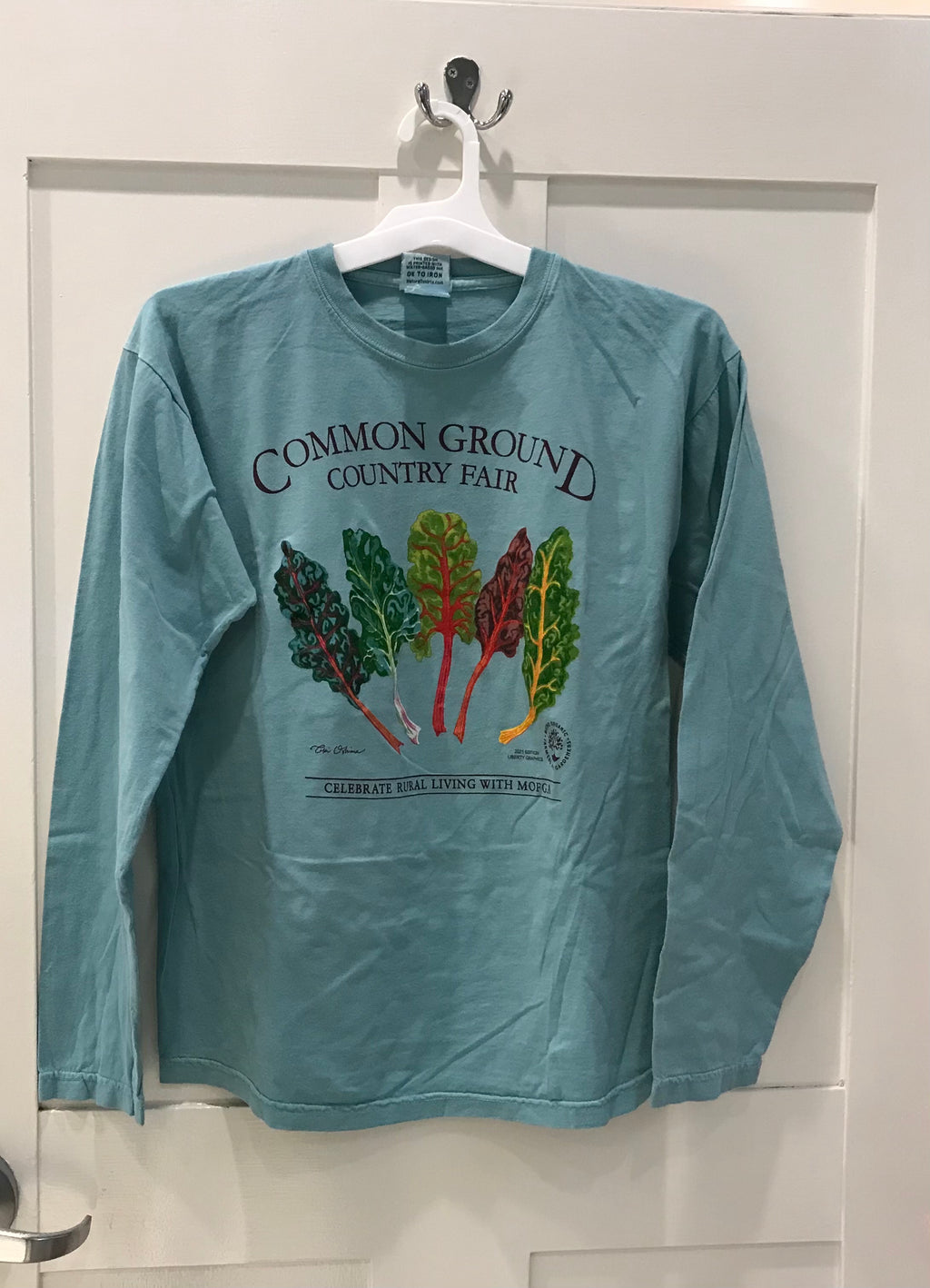 A light blue long sleeve shirt with artwork in the center depicting a colorful splay of five green, yellow, red, and purple chard. Black text around art reads “Common Ground Country Fair” and  “Celebrate Rural Living with MOFGA”. A black MOFGA logo is in the bottom right corner. 