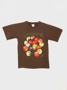 A navy shirt with artwork in the center depicting a cluster of 16 different apples. Each apple is small along the sides indicating the variety name and a Maine county. Lime green text around art reads “Common Ground Country Fair” and  “Celebrate Rural Living with MOFGA”. A lime green MOFGA logo is in the bottom right corner. 